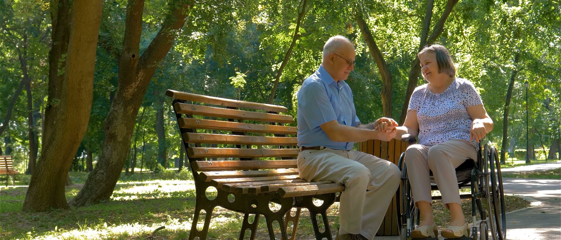 An older couple sit together on a sunlit park bench, holding hands and talking