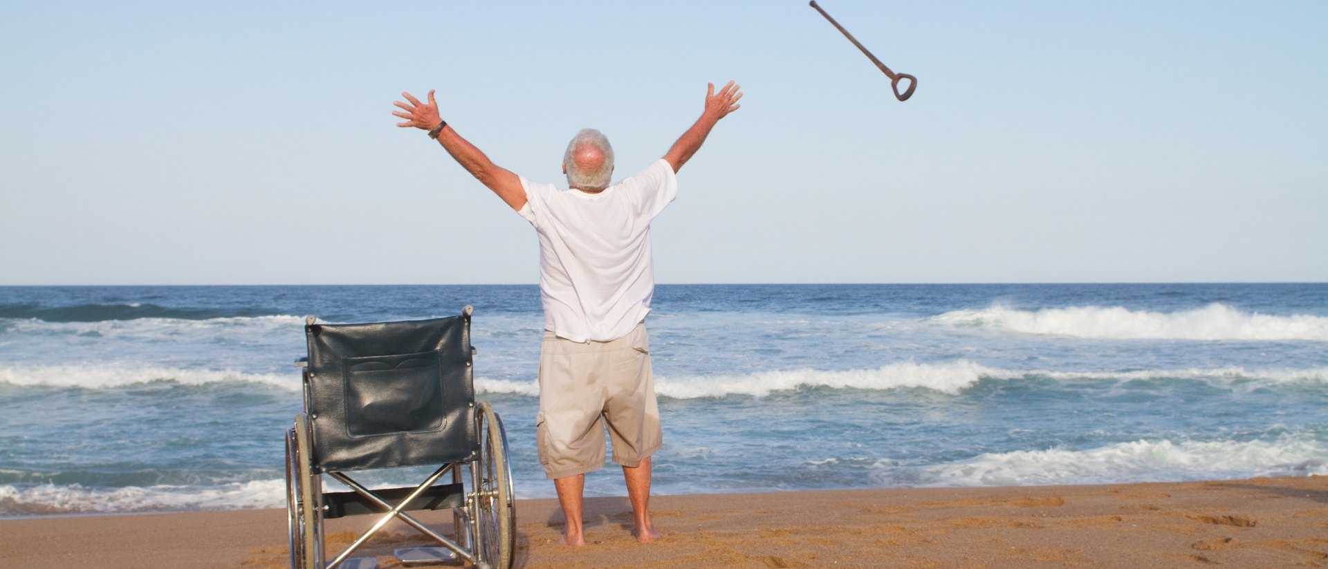 An older man stands at the water's edge with both arms raised, having just thrown his cane into the air, his folding wheelchair empty on the sand behind him