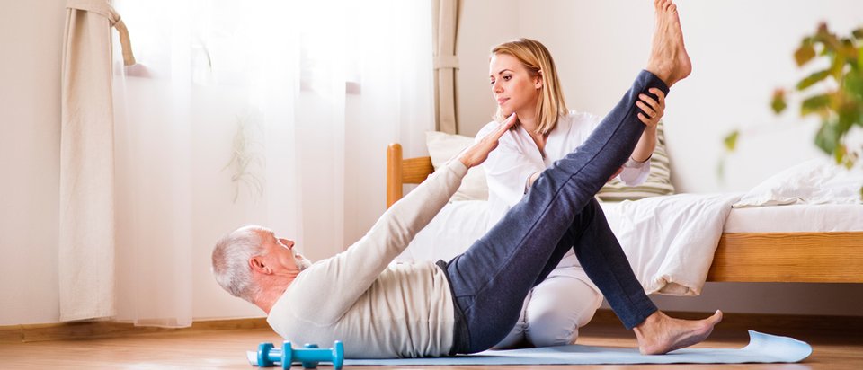 An older man lies on an exercise mat raising one leg while a physical therapist supports his foot from the bedside