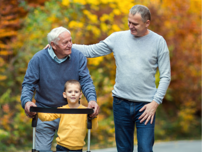Three generations walk a path together, a grandfather steadying himself on a rollator with his grandson riding in it and his son's hand on his shoulder