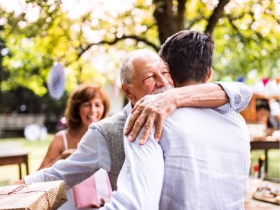 An older father and his adult son hold each other in a long embrace at a family gathering