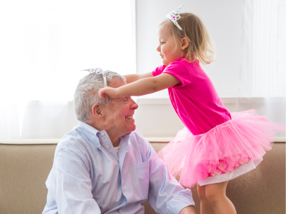 A great-grandfather sits on the sofa looking up at a toddler in a tutu and tiara who has reached up to pat his head