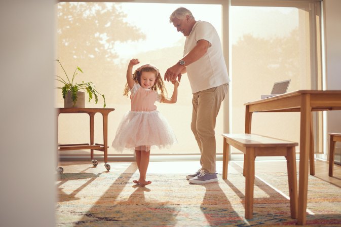 A grey-haired man dances hand in hand with a young girl in a pink tutu in a sunlit living room
