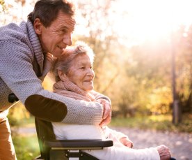 An older man in a grey cardigan leans down to embrace an elderly woman seated in a wheelchair, outdoors in autumn light