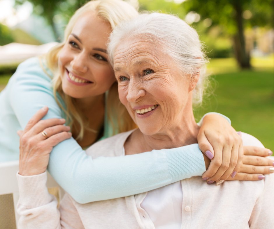 A smiling young woman hugs her white-haired grandmother from behind in a park