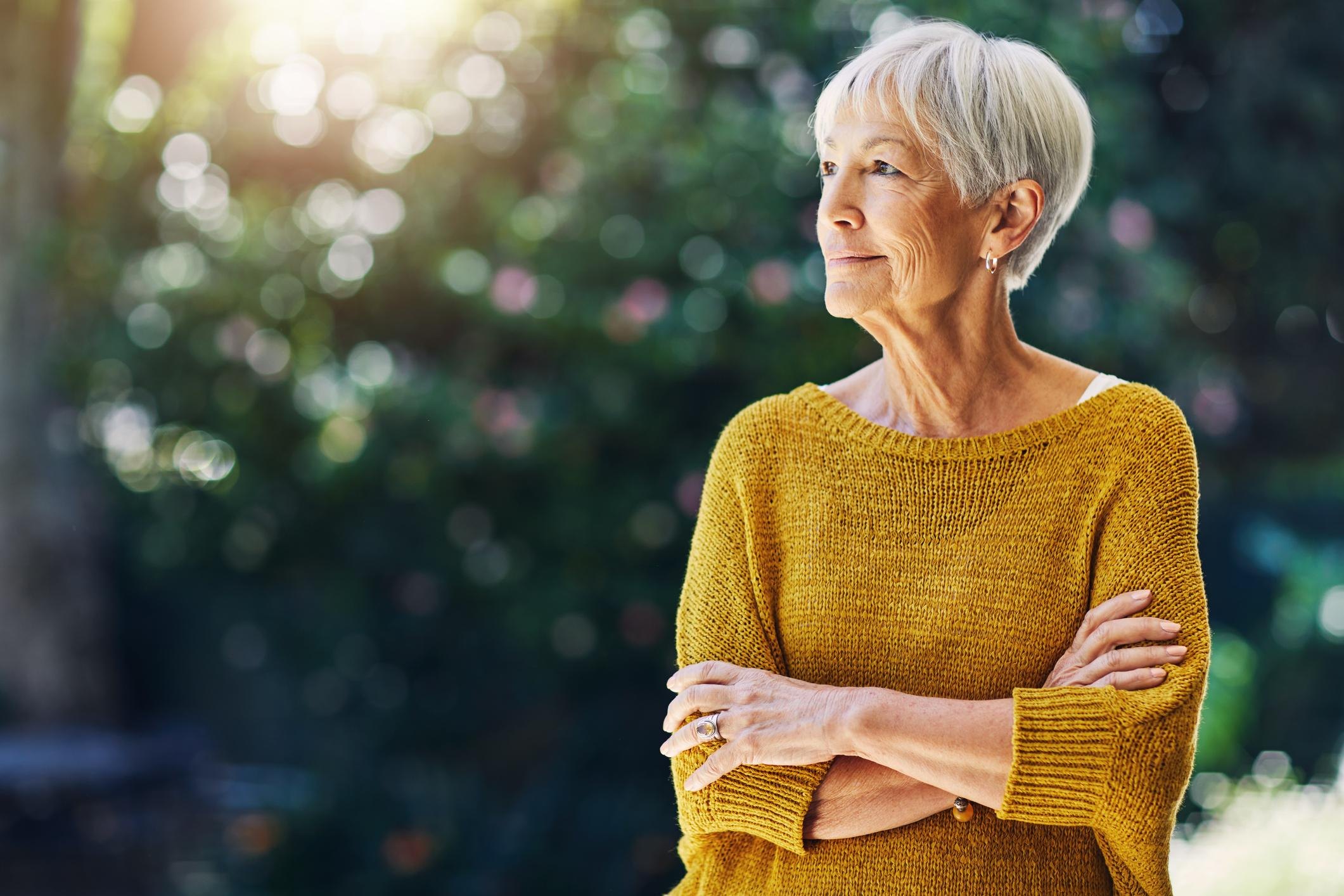 An older woman in a mustard sweater stands outdoors with her arms folded, looking into the distance