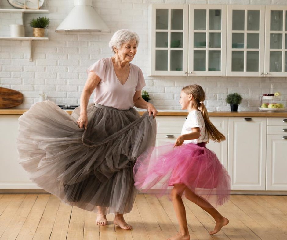 A grandmother in a grey tulle skirt dances barefoot in her kitchen with her granddaughter, who wears a pink tutu