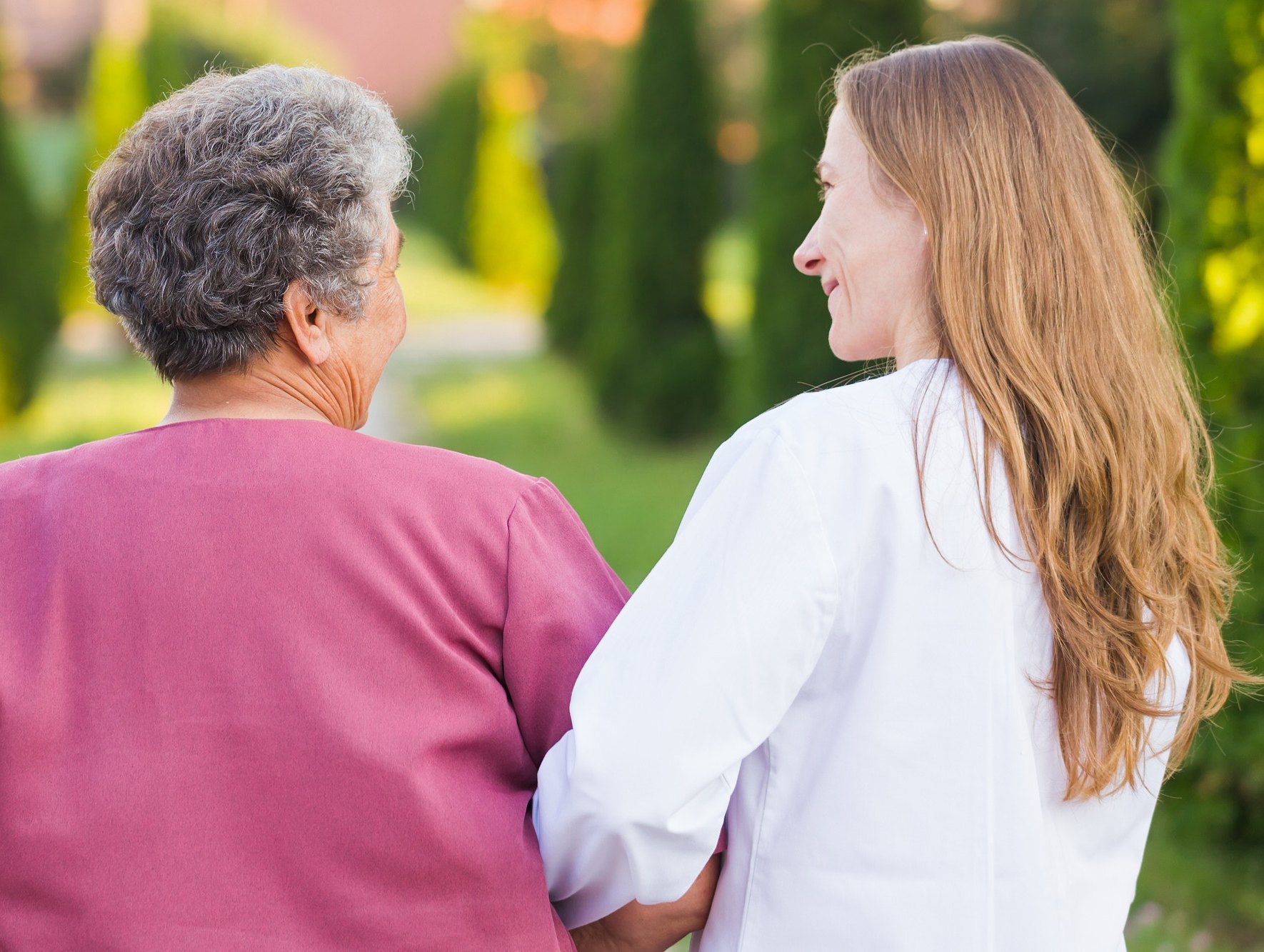 An older woman in a rose-colored top walks outdoors arm in arm with a nurse in a white coat