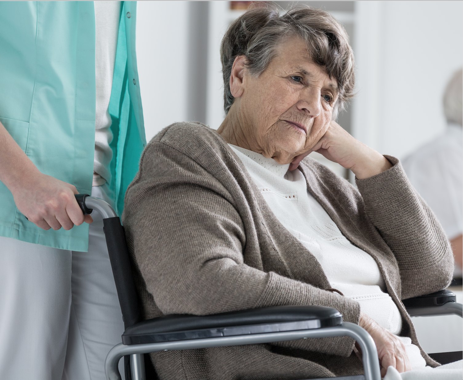 An older woman in a tan cardigan sits in a wheelchair, chin resting on her hand, while a caregiver in scrubs stands behind her