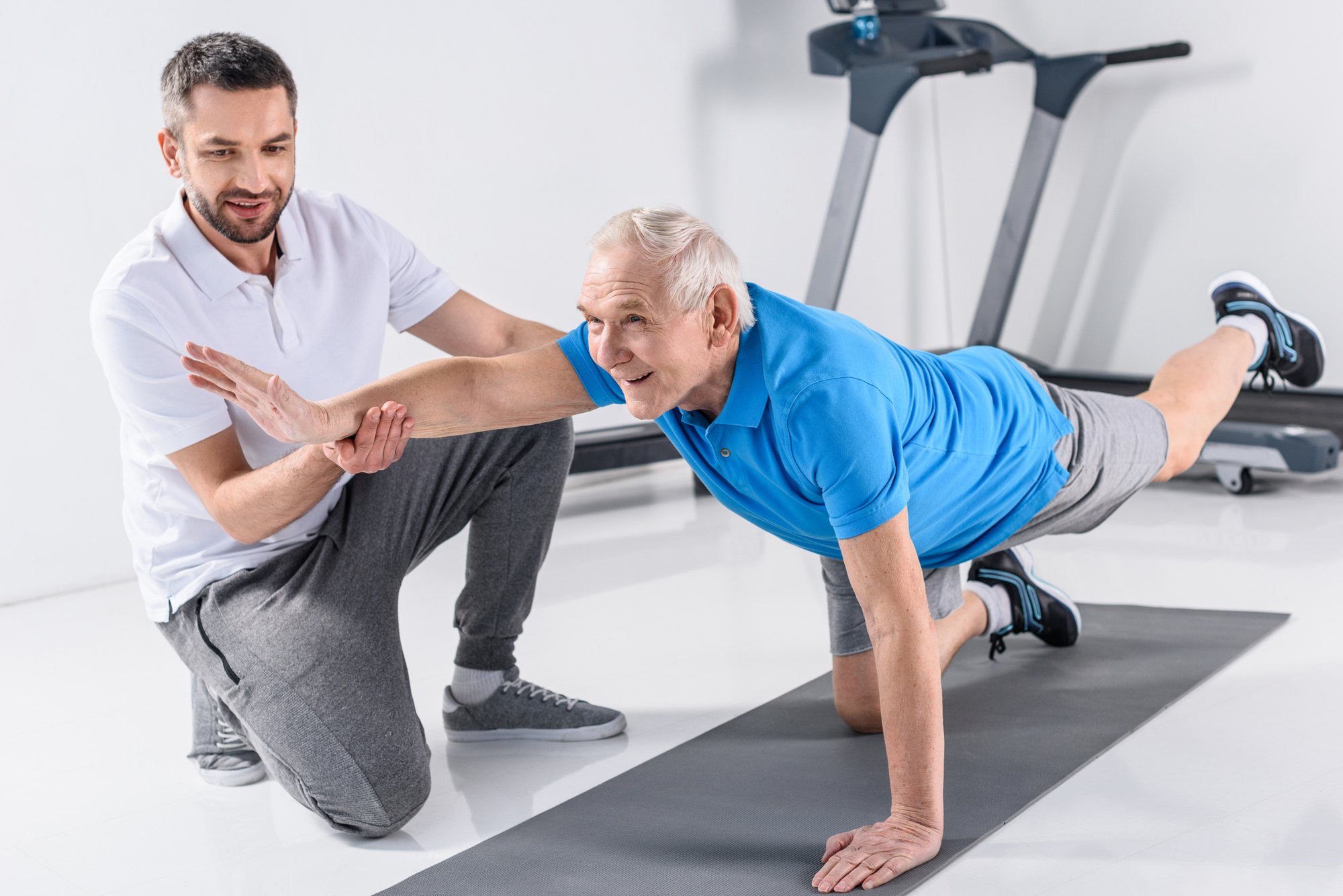 An older man in a blue polo holds a bird-dog exercise on a mat while a trainer supports his arm