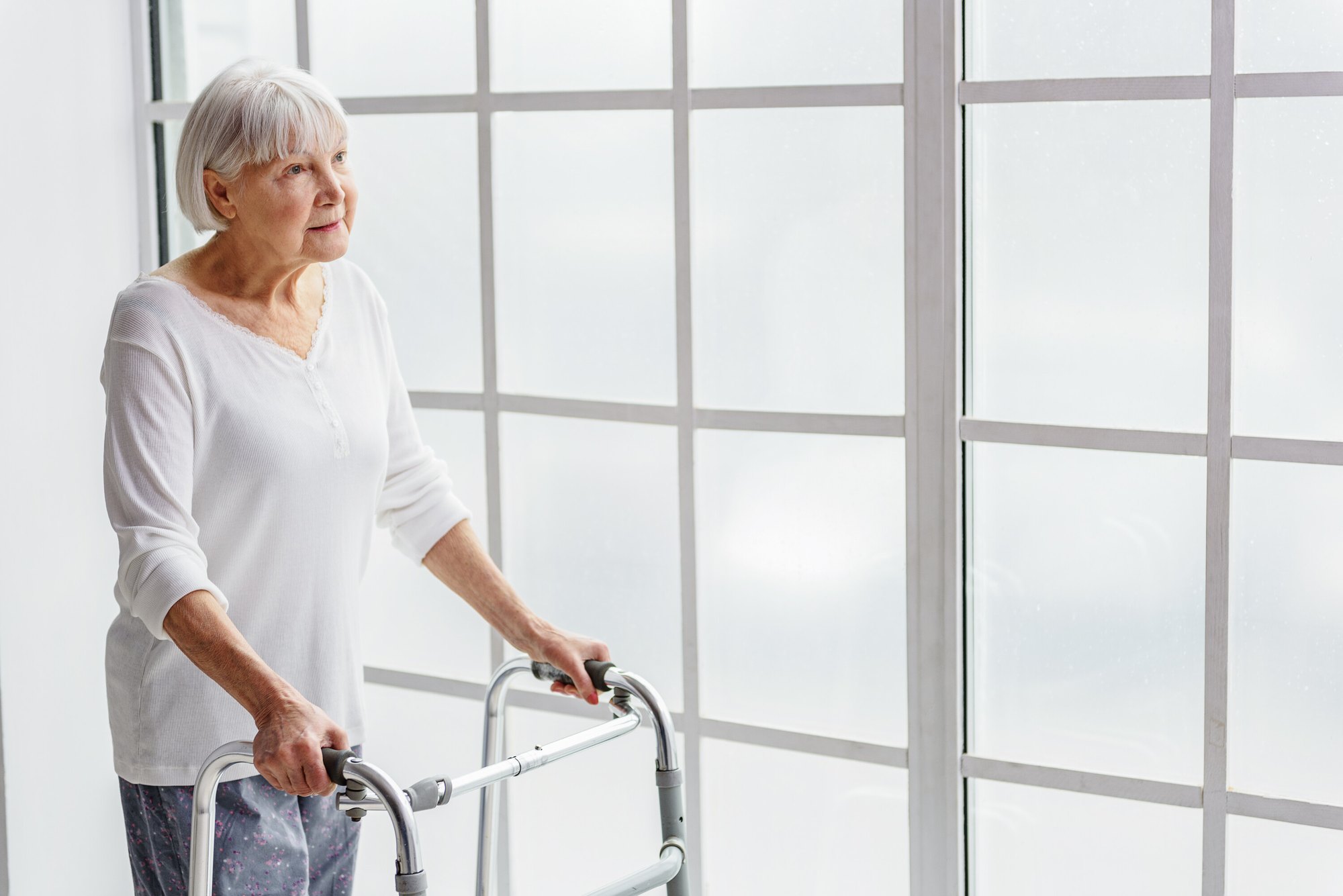 An older adult stands at a window with a walker, light coming in from outside