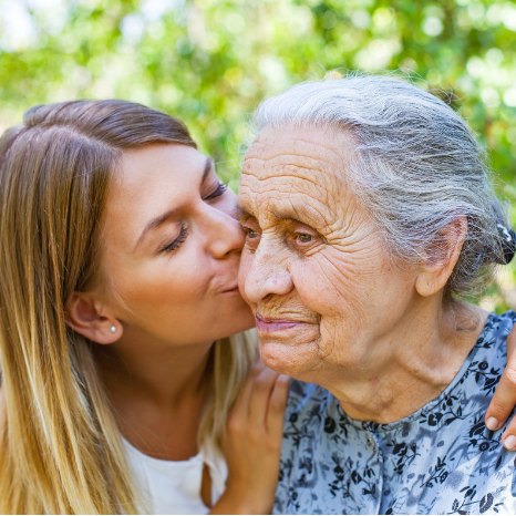 A young woman kisses her grandmother on the cheek outdoors under green trees
