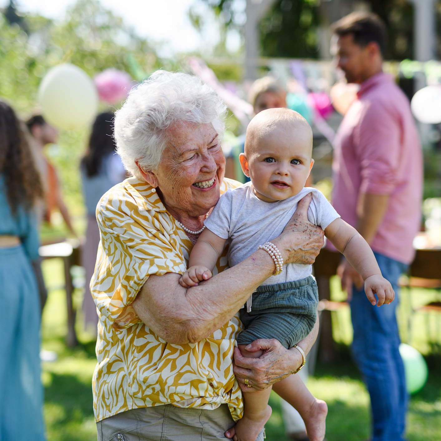 A laughing white-haired grandmother holds a baby at an outdoor garden party strung with balloons and bunting