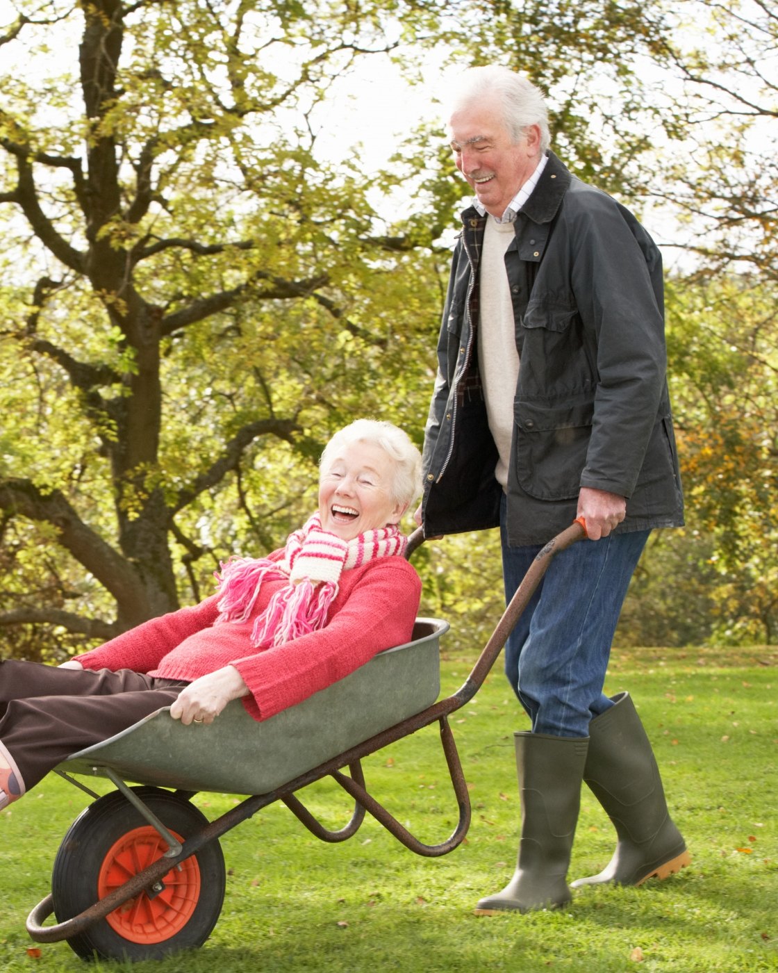 An older man laughs as he pushes his wife across the lawn in a wheelbarrow, her wellington boots kicked up in the air
