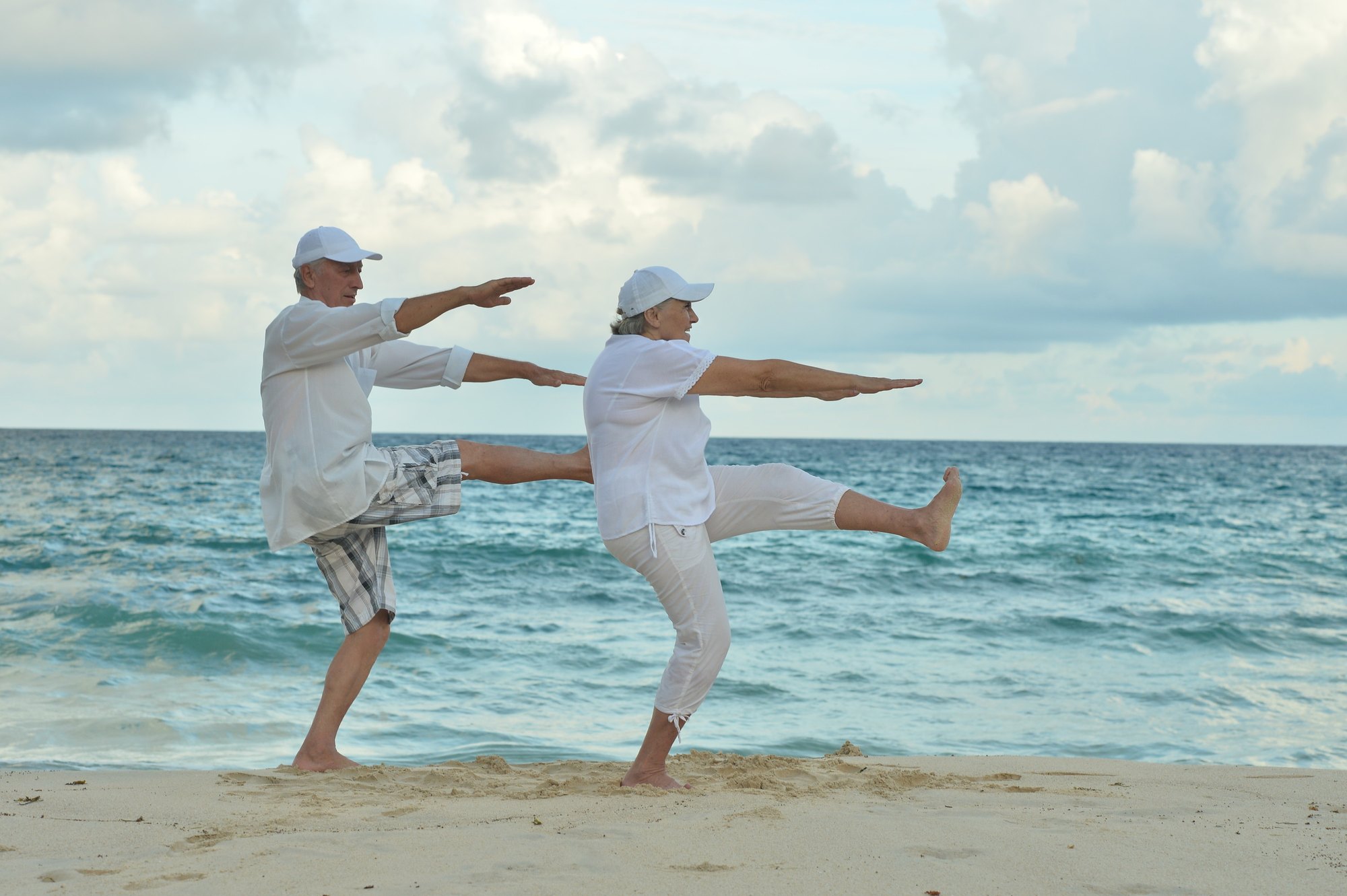 An older couple exercise on a beach, each balancing on one leg with arms outstretched