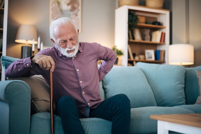 An older man with a white beard winces and holds his lower back while sitting on a sofa, one hand on his cane