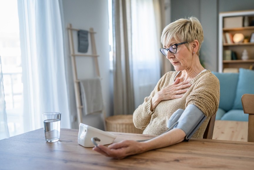 An older woman in glasses sits at a table with a blood pressure cuff on her arm, one hand resting on her chest