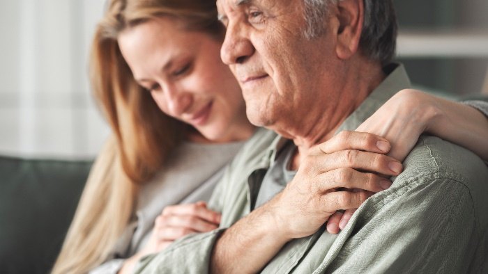 An adult daughter wraps her arms around her father from behind as they sit together on a sofa