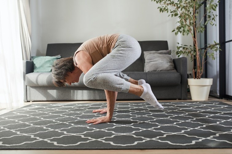 An older woman holds a crow pose, balancing on her hands on a patterned rug in her living room