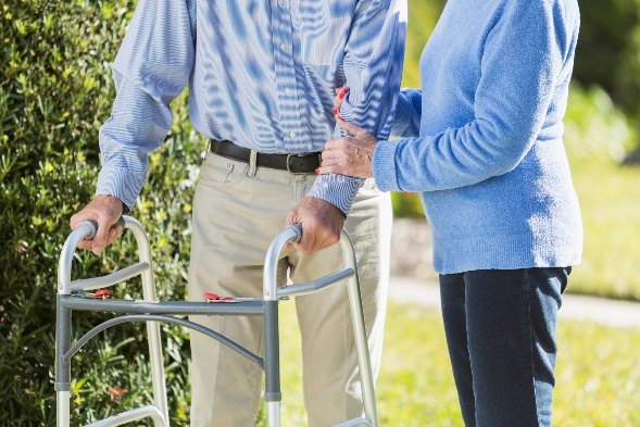 A woman in a blue sweater steadies the arm of an older man walking outdoors with a walker