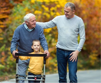 A grandfather with a walker, his grandson standing inside the frame and his adult son with a hand on his shoulder, on an autumn path