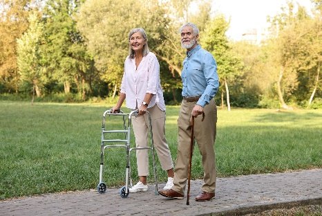An older couple walk side by side on a sunny park path, she with a wheeled walker and he with a cane