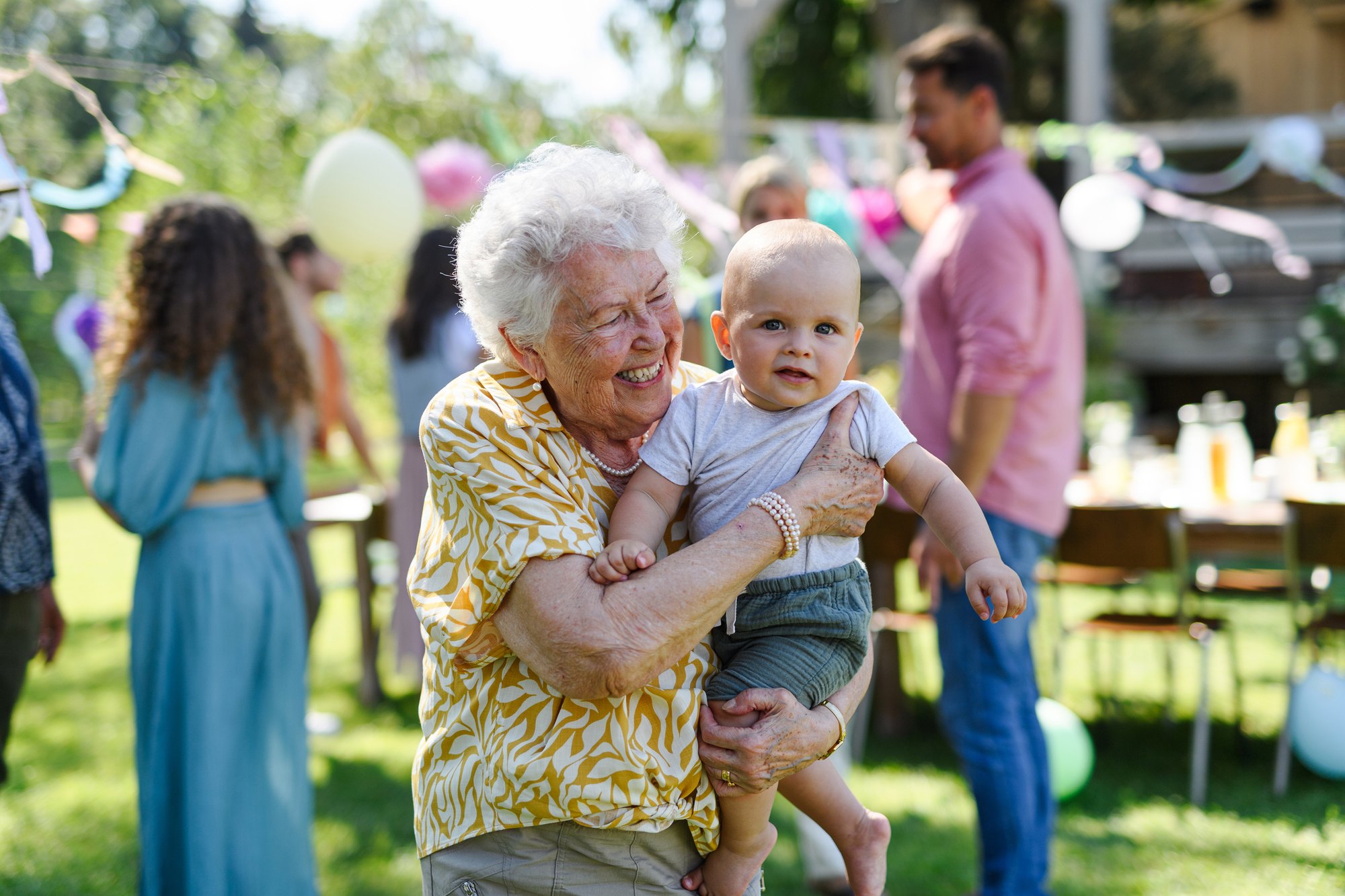 A grandmother holds a toddler at a garden party, balloons and family behind her