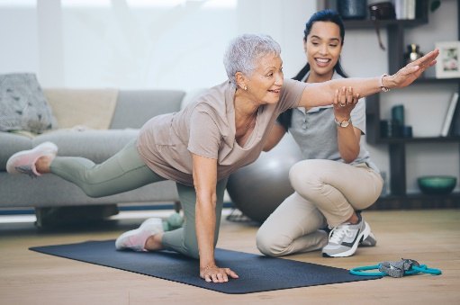 An older woman holds a bird-dog exercise on a mat at home while her therapist supports her outstretched arm