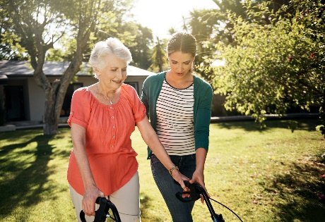 A caregiver walks beside an older woman using a walker in a sunny backyard