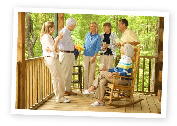 A multi-generational family talks and laughs together on a wooden porch surrounded by trees