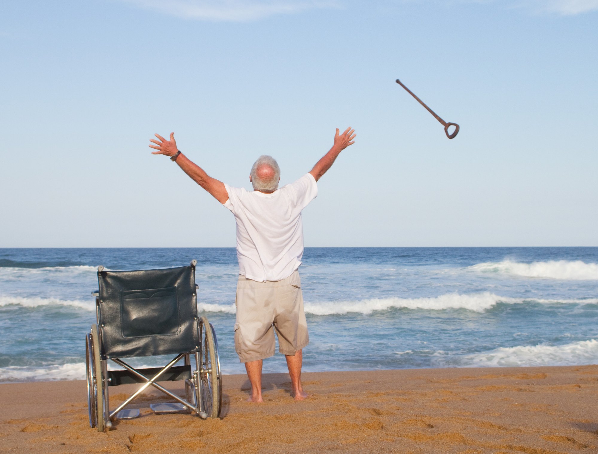 An older man stands barefoot on a beach with both arms raised, his empty wheelchair beside him and his cane flying through the air