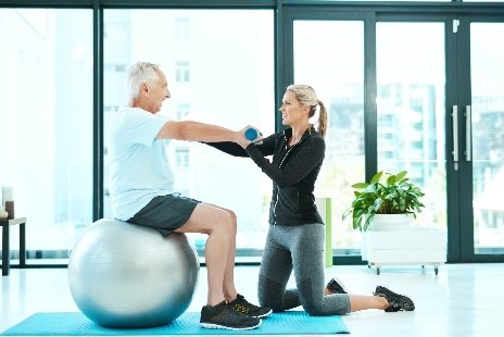 An older man seated on a stability ball works with his therapist in a bright studio