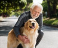 An older man crouches on a path to hug his golden retriever