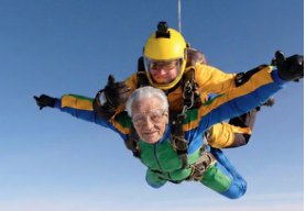 An older man skydives in tandem with an instructor against a blue sky
