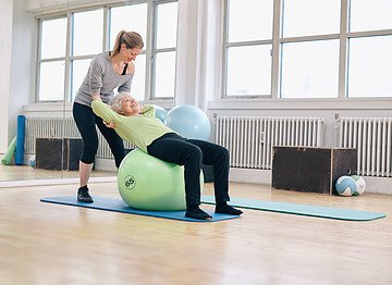 A trainer supports an older woman exercising on a large green ball