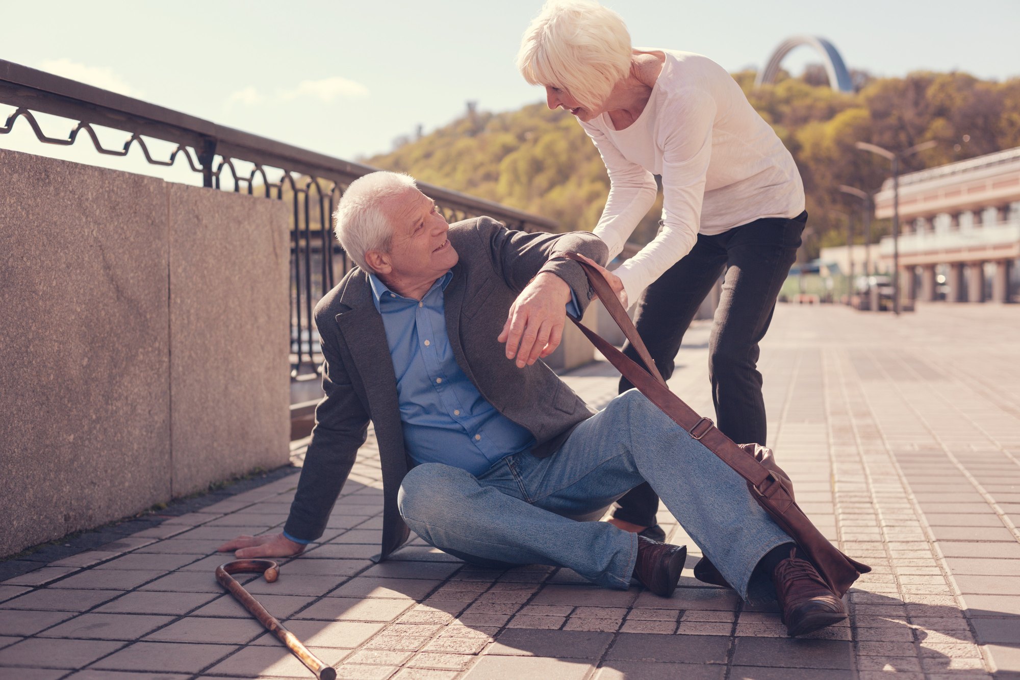 An older man sits on the pavement after a fall while a woman leans down to help him