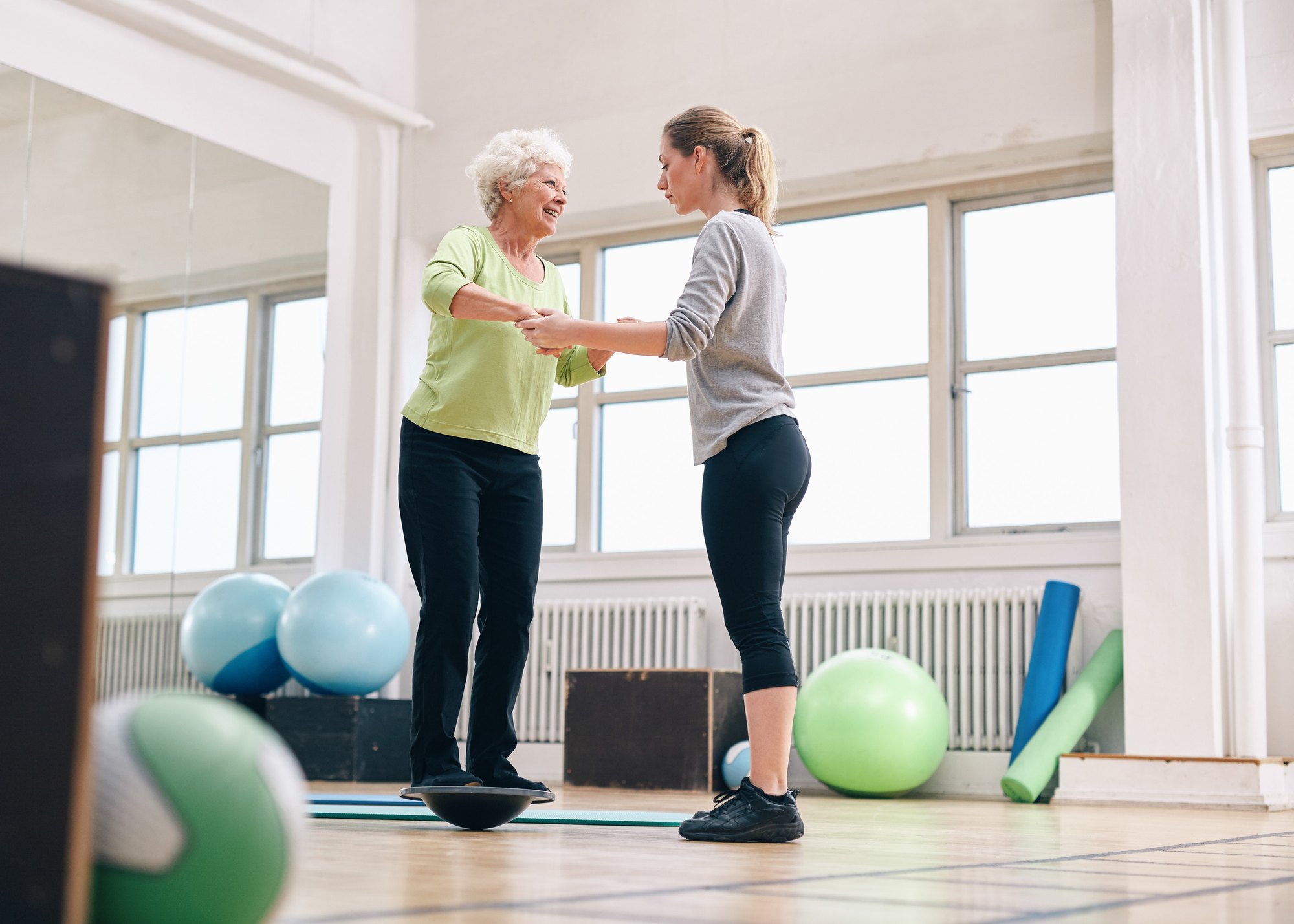 An older woman balances on a wobble board while a trainer holds her hands