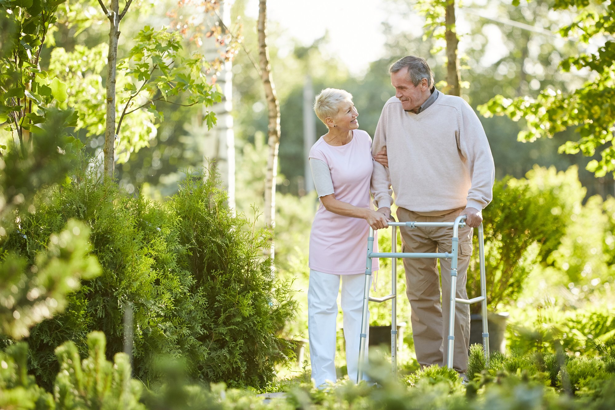 An older man walks with his walker through a sunny garden while a woman steadies his arm