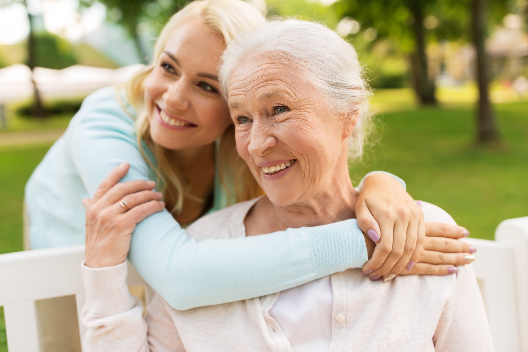 A younger woman hugs an older woman from behind as they share a park bench