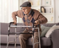 An older man in a flat cap and cardigan pushes up from a sofa, gripping his walker