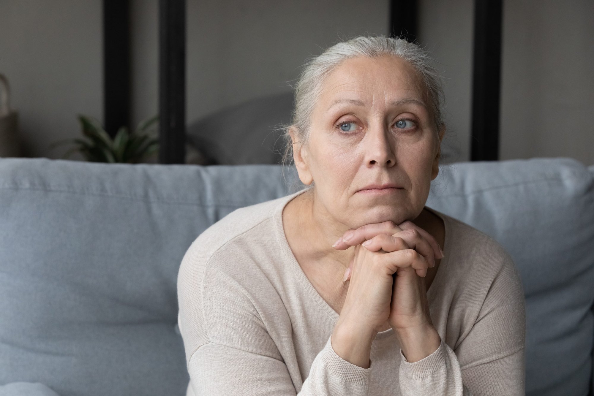 A woman sits on a couch with her hands clasped, looking thoughtful