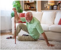 An older man sits on a living-room rug gripping his cane