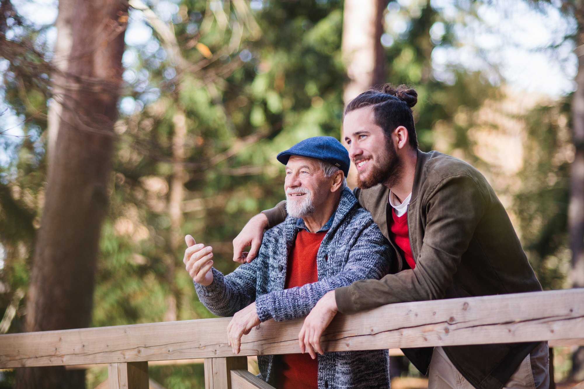 An older man and a younger man lean on a wooden railing in the woods, talking and smiling