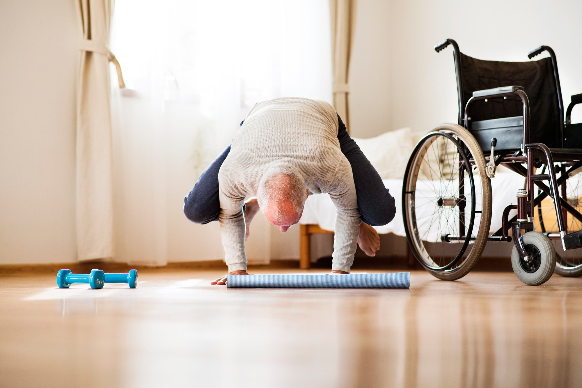 An older man holds a crow yoga balance on a mat at home, his wheelchair parked beside him